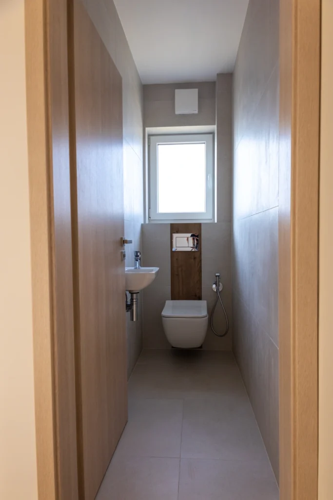 Toaleta v patře rodinného domu RD Kostelec nad Labem s moderním závěsným WC, malým umyvadlem a oknem pro denní světlo; interiér laděný do světlých neutrálních tónů s dřevěnými a keramickými prvky působí čistě a minimalisticky.
