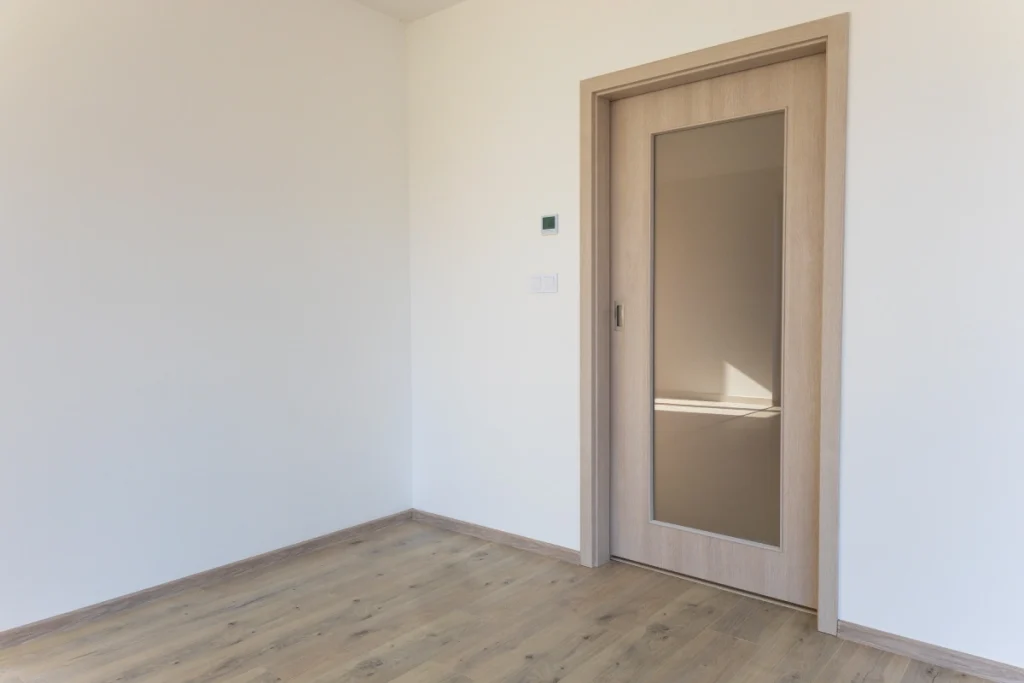 Detailní pohled na část obývacího pokoje v rodinném domě; světlá dřevěná podlaha, bílé stěny a posuvné dveře s matným sklem v dřevěném rámu dodávají prostoru moderní, minimalistický vzhled.