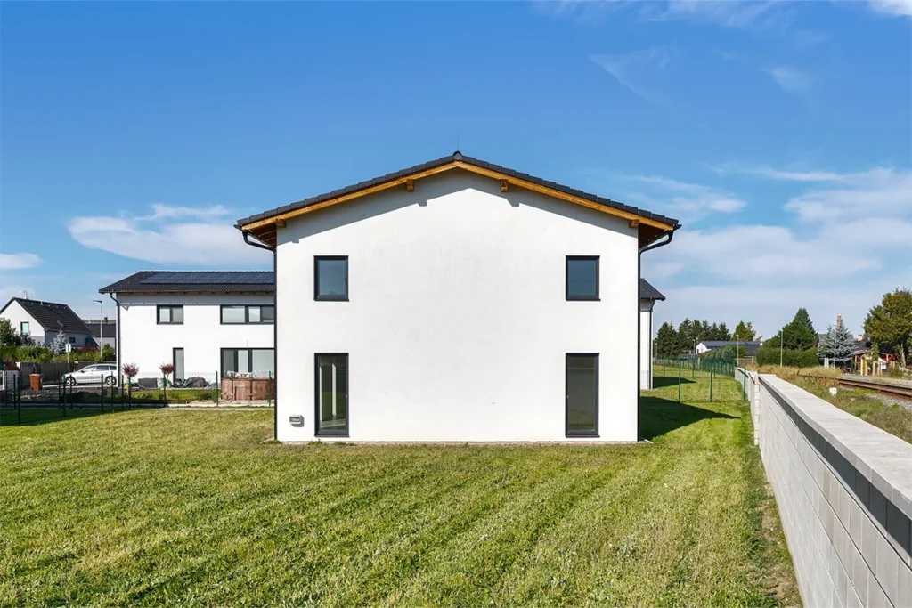Moderní dvoupodlažní rodinný dům s bílou a cihlovou fasádou, velkými okny a oplocenou zahradou.
